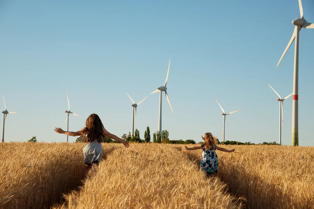 Raparigas correm por um campo na direção das turbinas eólicas.