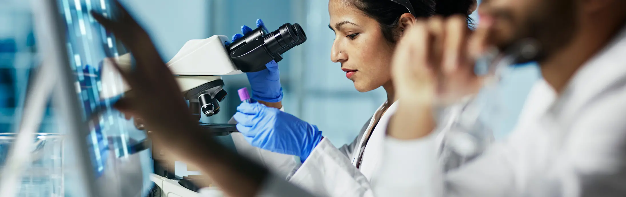 um laboratório com uma mulher em segundo plano sentada em frente a um microscópio, a verificar uma amostra e em primeiro plano um homem com barba a mexer no ecrã de um computador