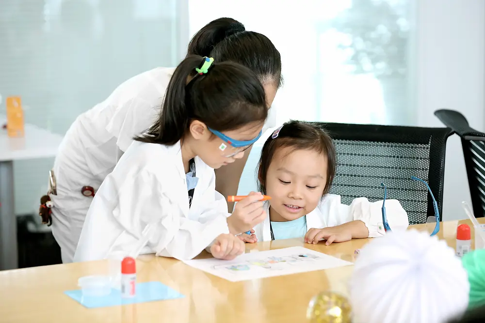 Duas crianças e uma mulher com uma bata de investigação pintam um quadro numa mesa.