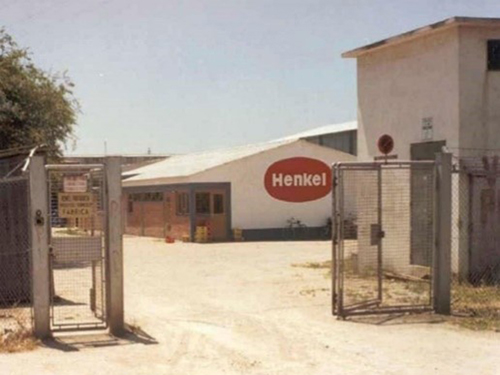 Entrada da fábrica de Adesivos para as indústrias têxteis onde você pode ver um prédio branco com um logotipo vermelho