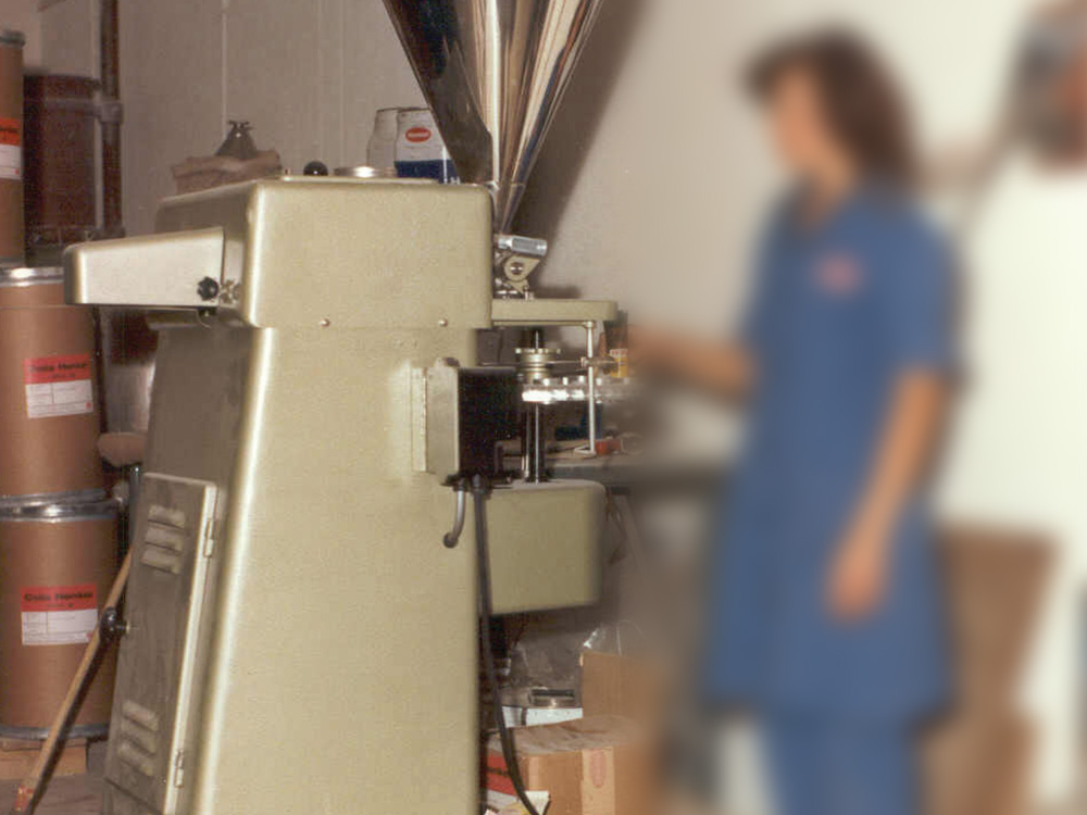 Máquina industrial bege operada por uma mulher turva de casaco azul e calça comprida.