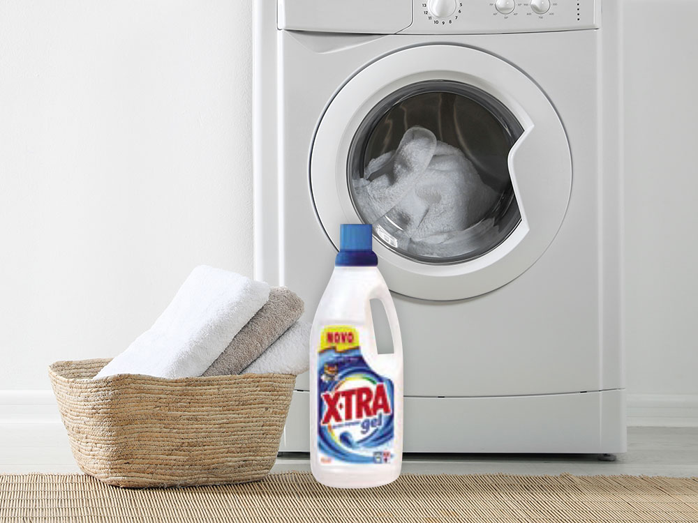 Detergente X-tra ao lado da roupa branca e da máquina de lavar.
