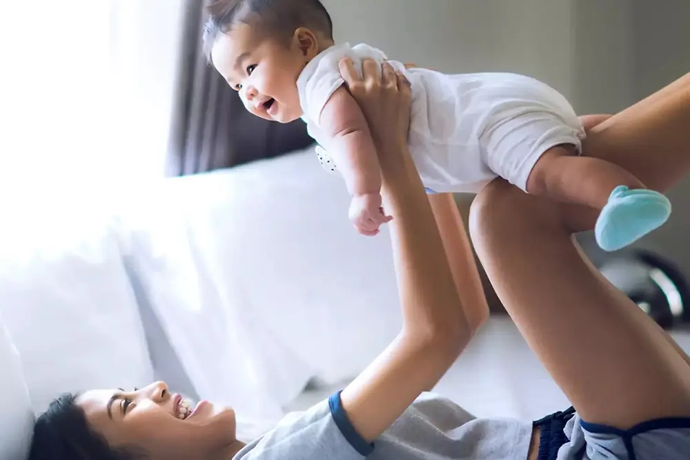 Mãe levanta-se e brinca com um bebé sorridente dentro de casa, num ambiente iluminado.