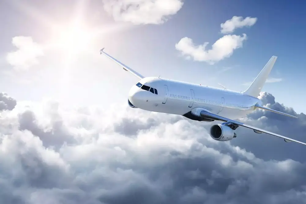 Um avião de passageiros branco voa acima de nuvens suaves em um céu brilhante, com luz solar vindo do canto superior esquerdo.