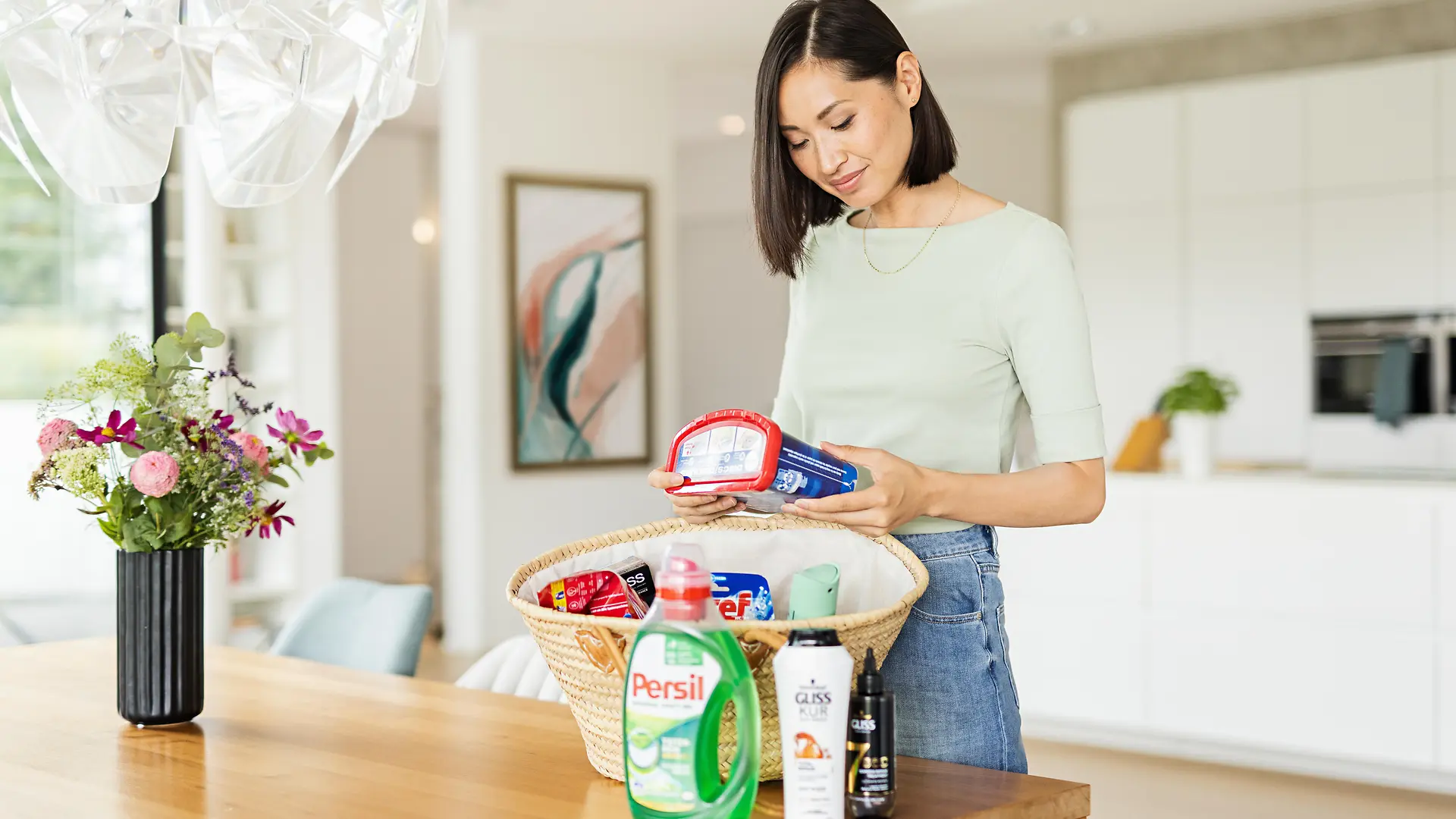 Mulher com um cesto em cima de uma mesa cheia de produtos Henkel, tais como: Persil, Gliss, Bref, Sonasol e mais. 