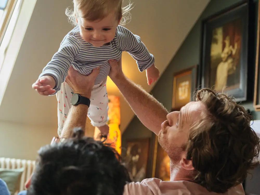 Dois pais a segurar o seu bebé e a sorrir em casa, simbolizando a inclusão e a política de licença parental da Henkel, favorável às famílias.
