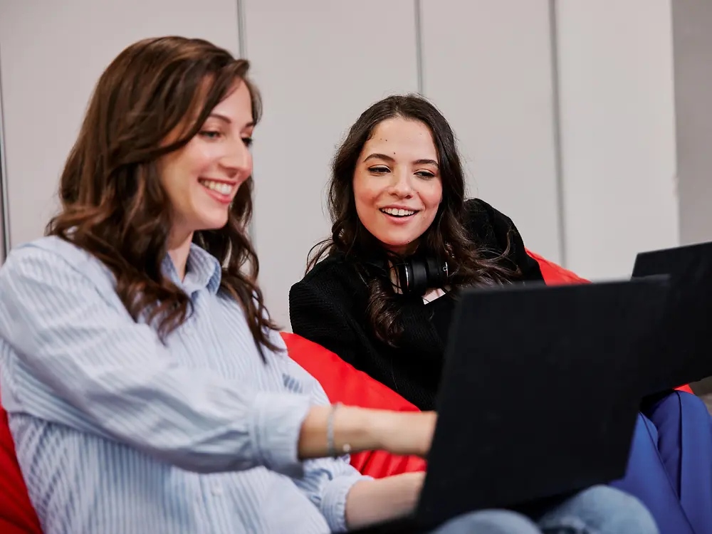 Duas pessoas sentadas em pufes coloridos num escritório moderno, cada uma a usar um portátil. Parecem estar a discutir os benefícios para colaboradores da Henkel enquanto trabalham em colaboração.