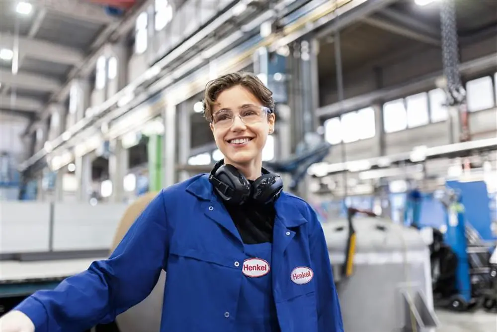 Colaborador da Henkel a sorrir, usando uma bata de trabalho azul e auscultadores de segurança, de pé numa unidade de produção industrial, representando experiência e inovação na produção.