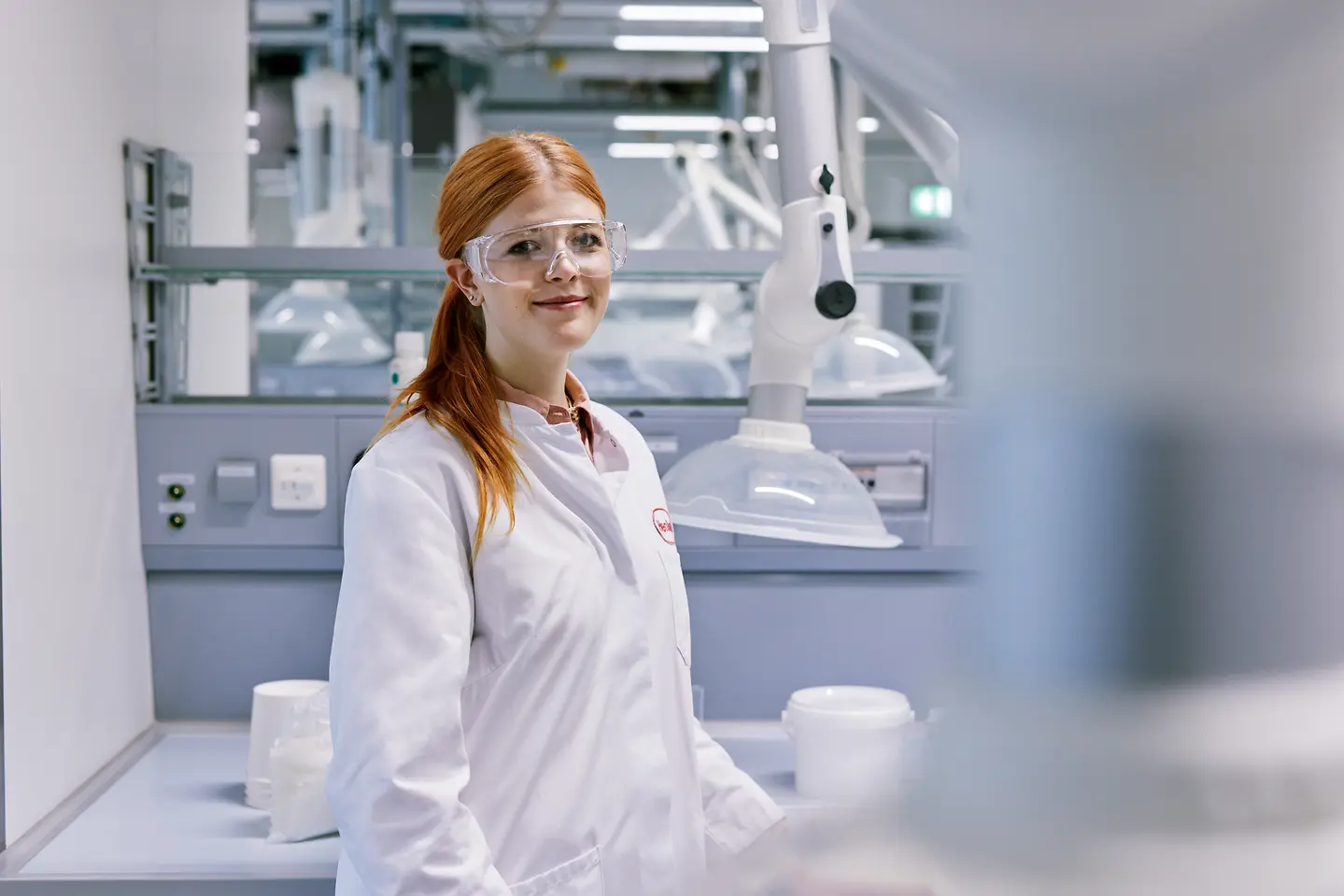 Recém-graduado a iniciar a sua carreira na Henkel, com uma bata branca, de pé num ambiente de laboratório moderno com equipamento técnico ao fundo.