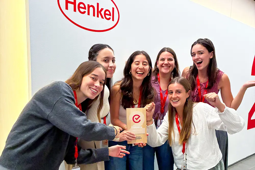 Grupo de seis pessoas de pé em frente a uma parede com o logótipo da Henkel. Usam cordões de identificação vermelhos e roupa casual, e estão reunidas de perto à volta de uma placa de prémio. O ambiente parece ser um espaço corporativo interior, com uma parede clara e pavimento cinzento. O grupo posa de forma entusiástica, destacando o prémio no centro.