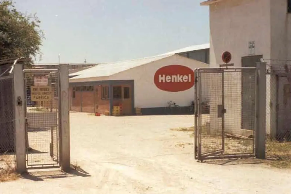 Entrada da fábrica da Henkel em Alverca, com portão metálico, edifício baixo com logótipo Henkel visível e uma área exterior pavimentada.