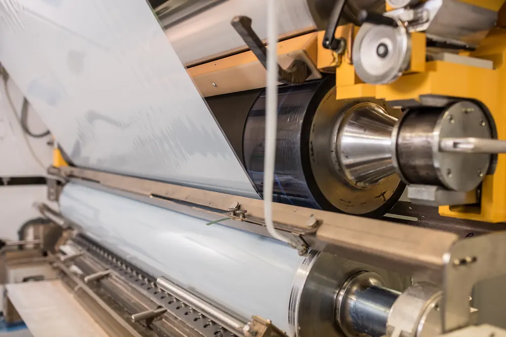 Máquina industrial a aplicar ou laminar película adesiva sobre um rolo, com cilindros metálicos em funcionamento numa linha de produção.