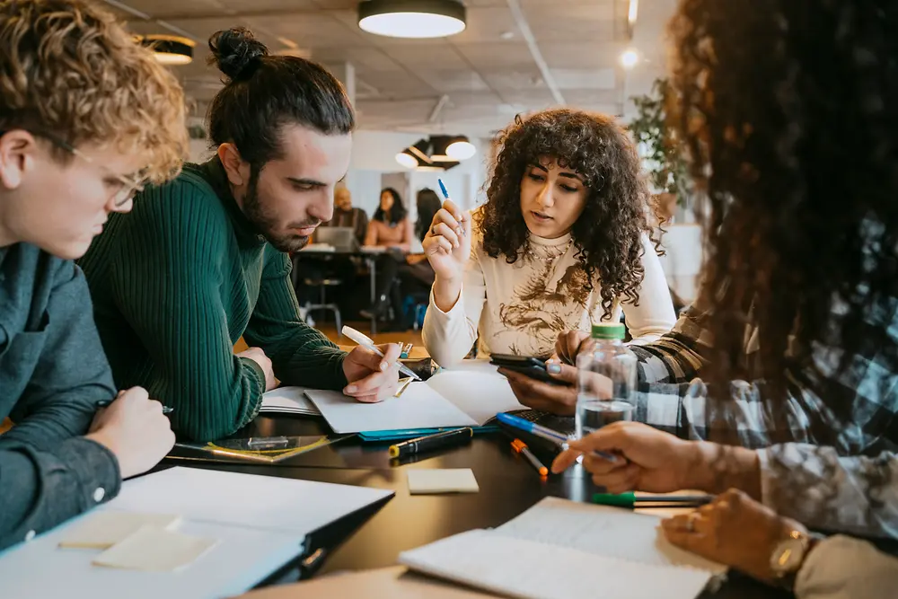 Um grupo de pessoas sentadas à volta de uma mesa num espaço de trabalho interior moderno, a colaborar em documentos, cadernos e dispositivos digitais. Várias canetas, folhas de papel e notas adesivas estão espalhadas pela mesa, enquanto o grupo participa numa discussão e toma notas.