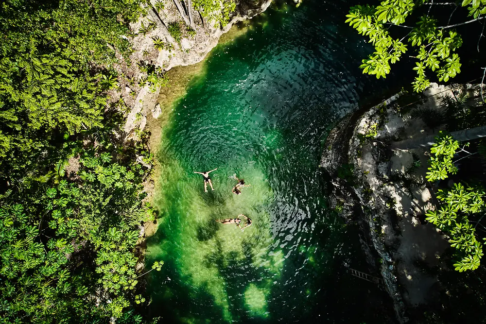Vista aérea de uma pequena piscina natural rodeada por densa vegetação verde, com três pessoas a flutuar na água límpida de cor esmeralda.