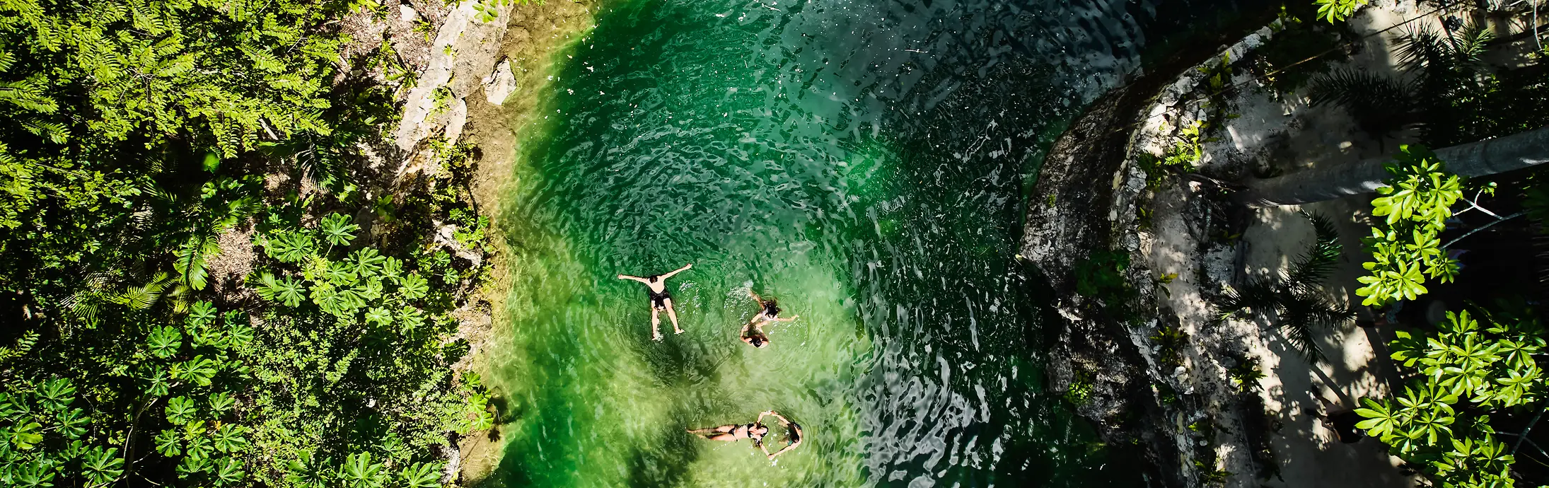 Vista aérea de uma pequena piscina natural rodeada por densa vegetação verde, com três pessoas a flutuar na água límpida de cor esmeralda.