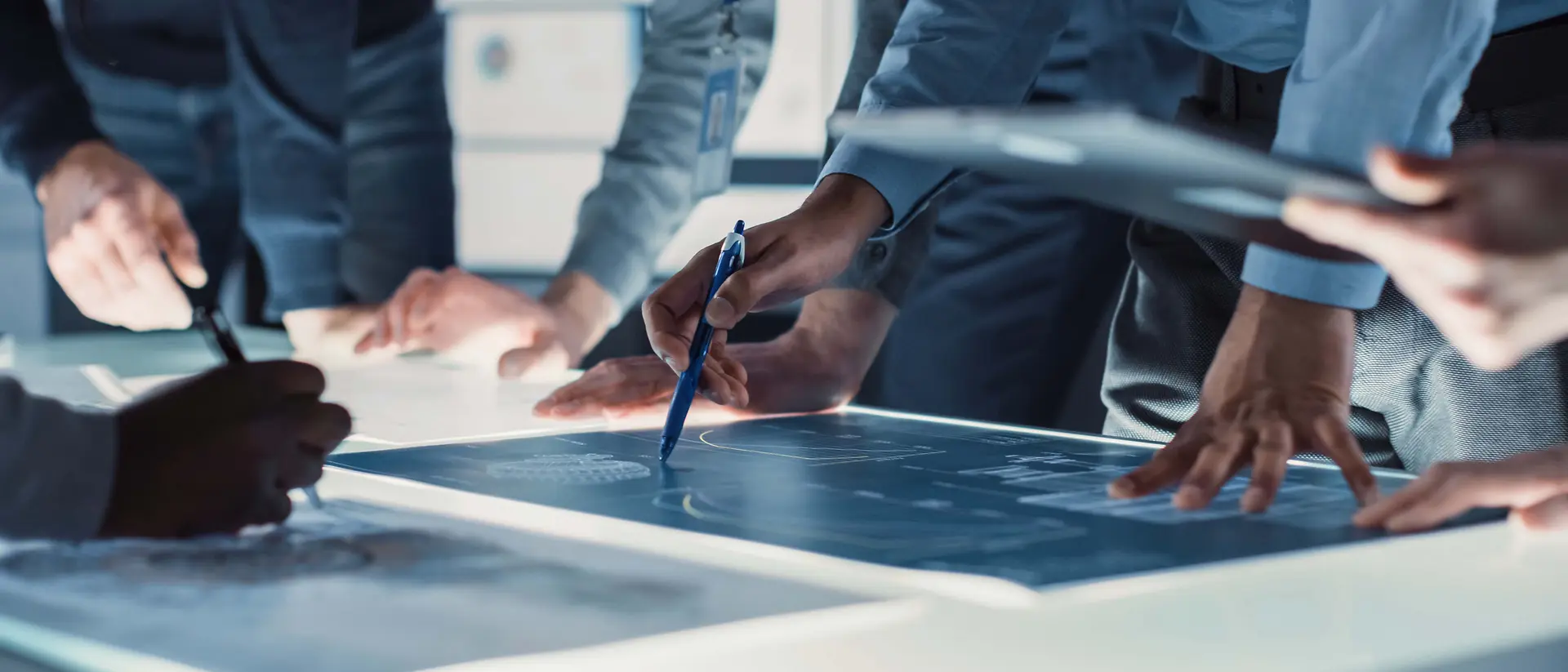 Imagem em grande plano de várias pessoas reunidas em torno de uma grande mesa iluminada, a analisar e a assinalar desenhos técnicos com canetas e dispositivos digitais.