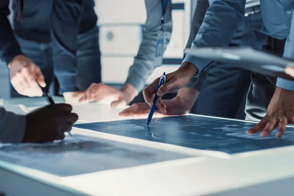 Imagem em grande plano de várias pessoas reunidas em torno de uma grande mesa iluminada, a analisar e a assinalar desenhos técnicos com canetas e dispositivos digitais.