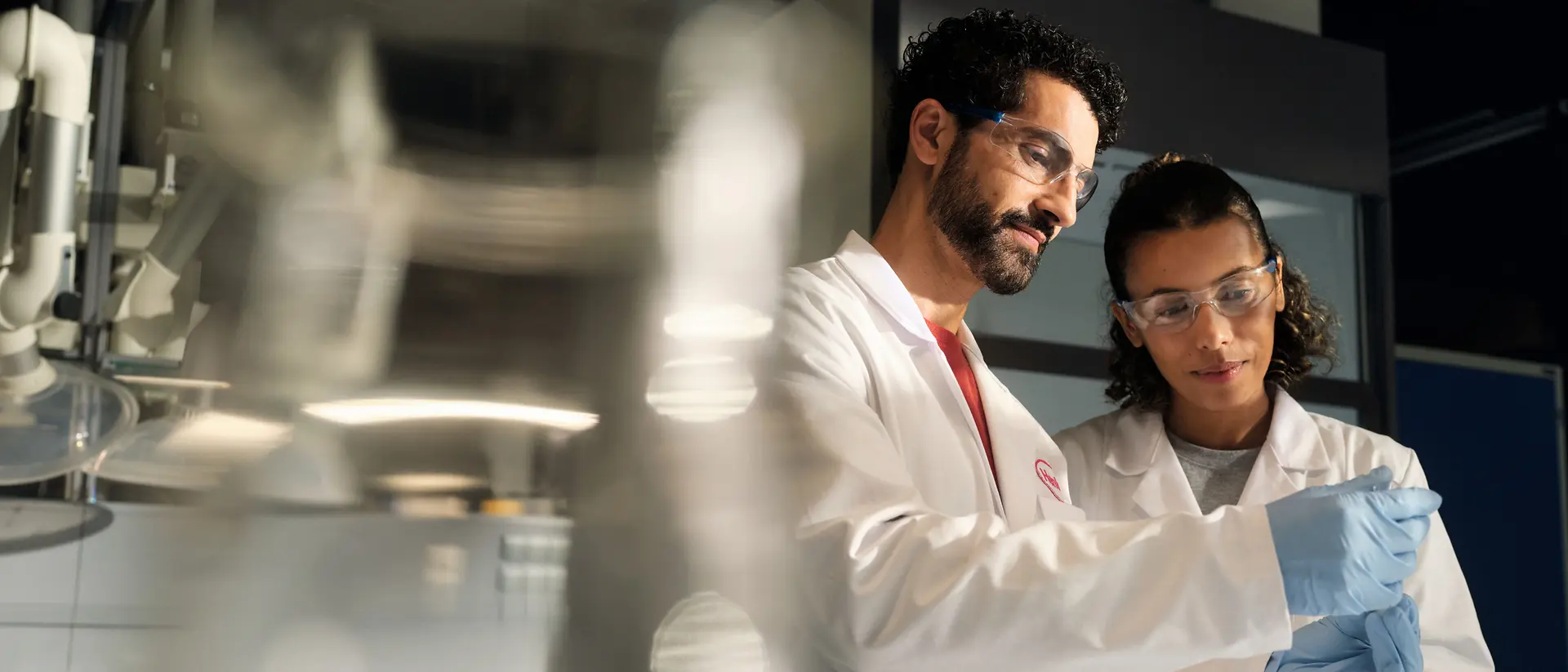 Dois cientistas, vestidos com batas brancas de laboratório, óculos de proteção e luvas, examinam juntos um pequeno recipiente com uma amostra num ambiente de laboratório.