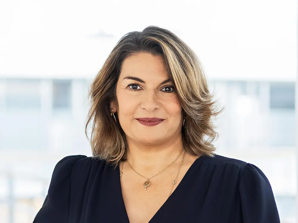 Retrato corporativo de Sylvie Nicol, diretiva da Henkel, com blusa azul-escura e cabelo castanho, posando num escritório luminoso.