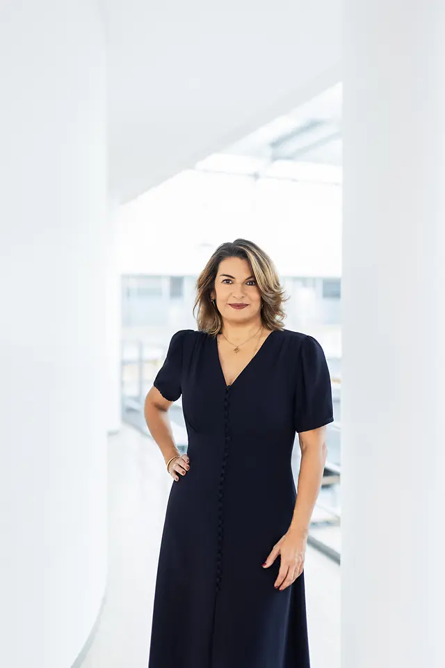 Retrato corporativo de Sylvie Nicol, diretiva da Henkel, com blusa azul-escura e cabelo castanho, posando num escritório luminoso.