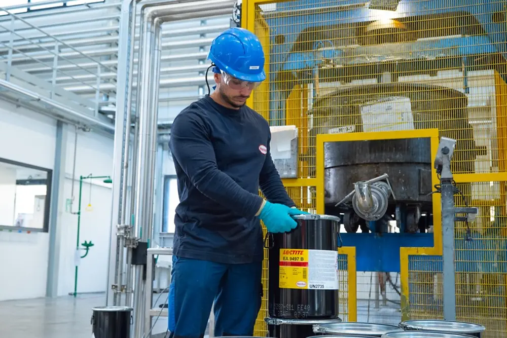 Ambiente industrial com um operário a manipular e a posicionar um recipiente de adesivo LOCTITE EA 9497 junto a maquinaria num contexto de produção.