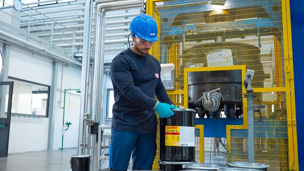 Ambiente industrial com um operário a manipular e a posicionar um recipiente de adesivo LOCTITE EA 9497 junto a maquinaria num contexto de produção.