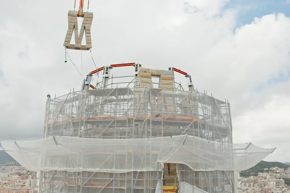 Processo de instalação de um painel na parte superior da torre de Jesus Cristo da Sagrada Família, com a estrutura em construção e uma grua a elevar o elemento.