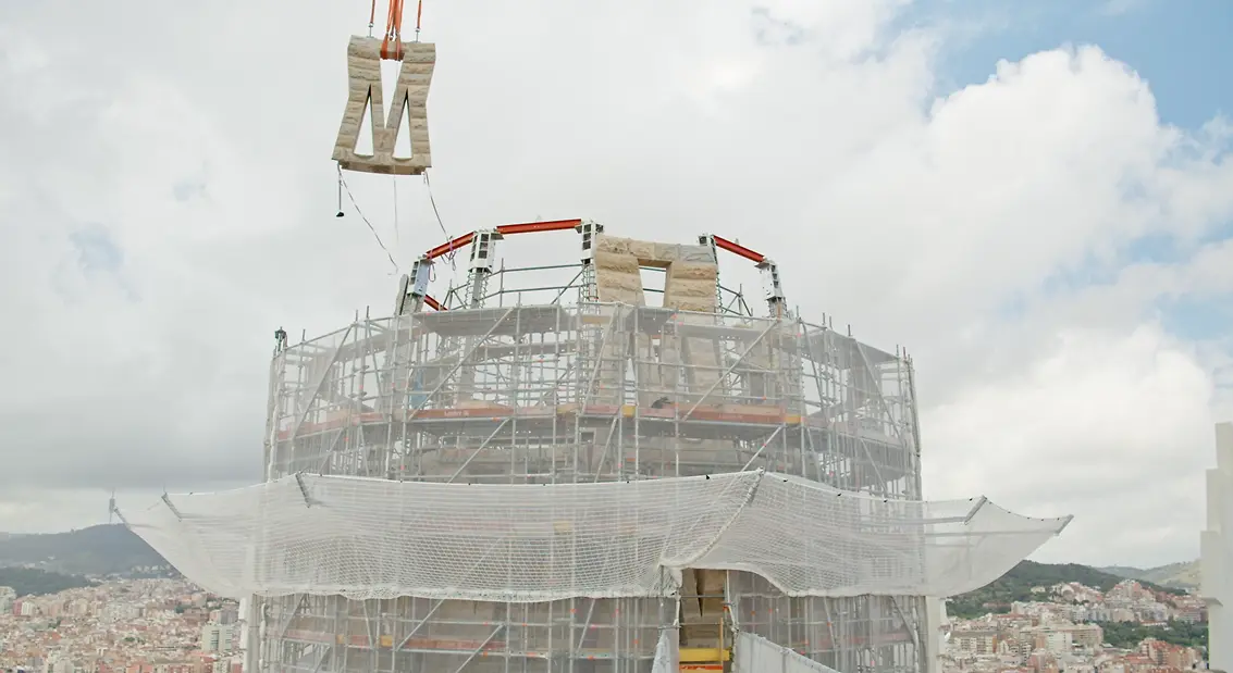 Processo de instalação de um painel na parte superior da torre de Jesus Cristo da Sagrada Família, com a estrutura em construção e uma grua a elevar o elemento.