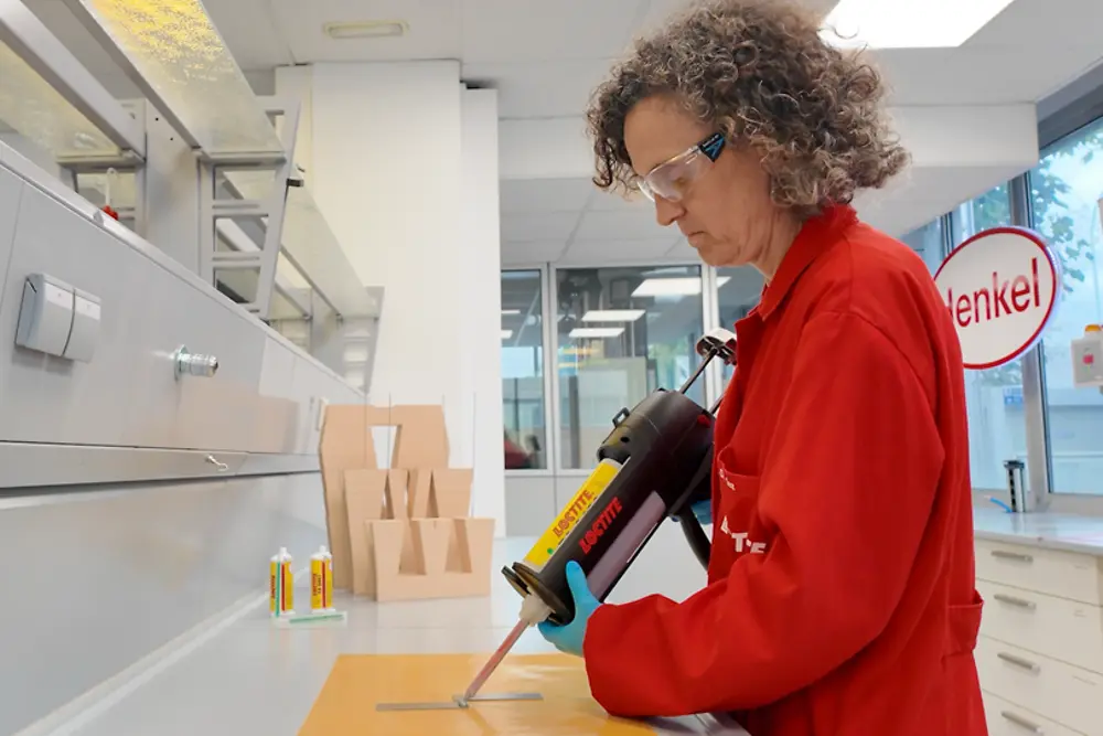 Ambiente de laboratório onde é aplicado adesivo com uma ferramenta de precisão sobre peças numa mesa de trabalho durante testes técnicos.