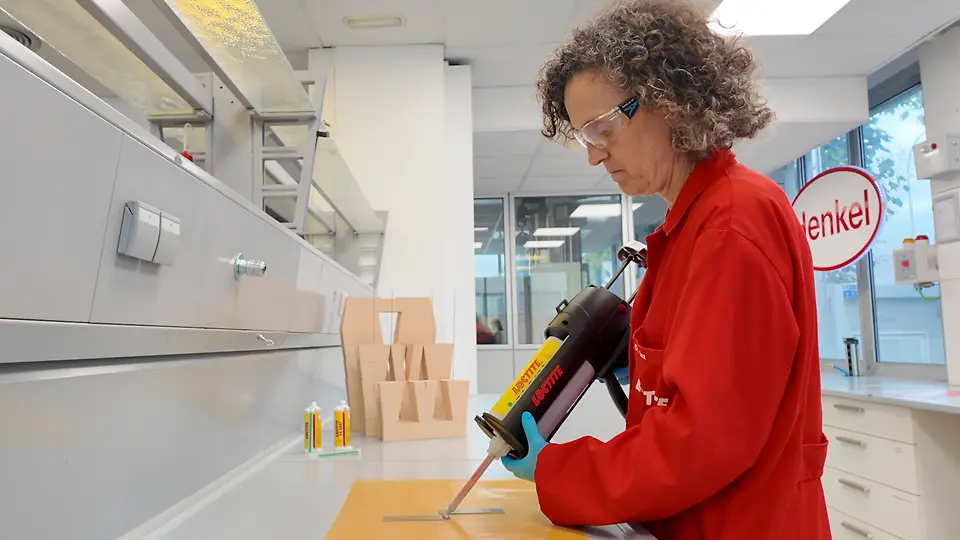 Ambiente de laboratório onde é aplicado adesivo com uma ferramenta de precisão sobre peças numa mesa de trabalho durante testes técnicos.