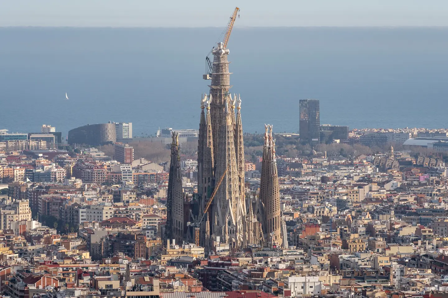 Vista geral da Sagrada Família, em Barcelona, com as torres centrais em construção e quatro pessoas em primeiro plano, em frente ao templo, numa praça urbana com peões e elementos de mobiliário urbano.