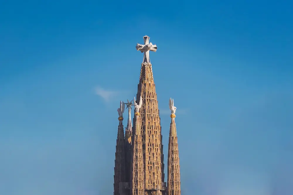 Vista vertical das torres centrais da Sagrada Família, destacando a torre principal coroada por uma cruz sobre um céu limpo.