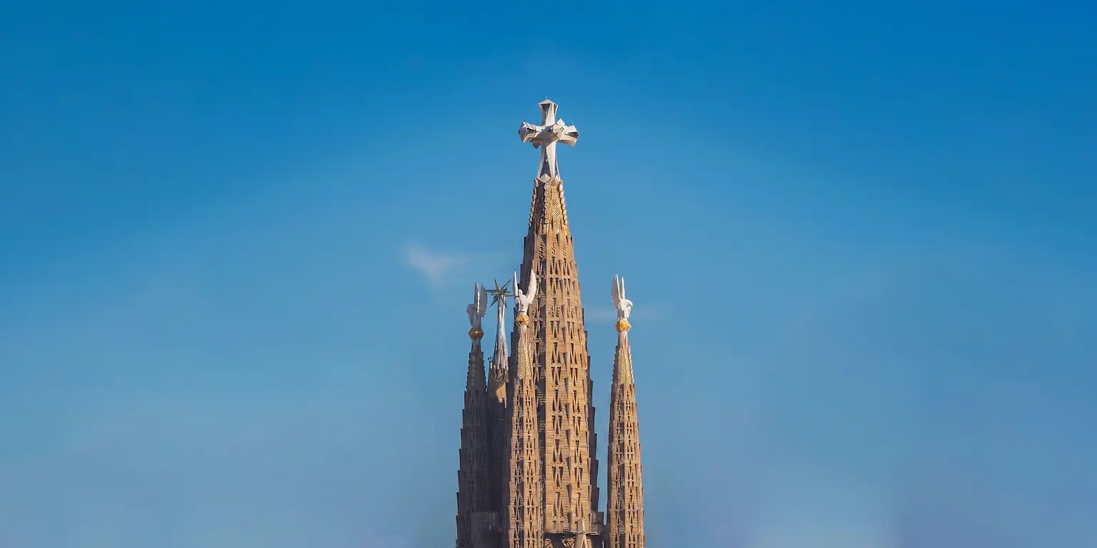 Vista vertical das torres centrais da Sagrada Família, destacando a torre principal coroada por uma cruz sobre um céu limpo.
