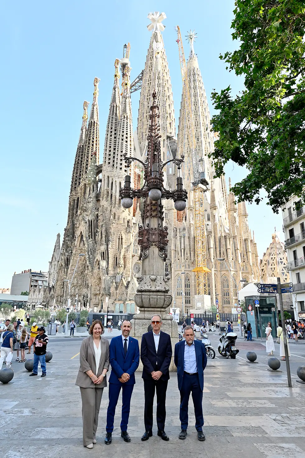 Vista geral da Sagrada Família em Barcelona, com as suas torres centrais em construção e quatro pessoas a posar em primeiro plano, em frente ao templo numa praça urbana com peões e elementos de mobiliário urbano.