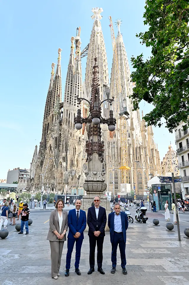 Vista geral da Sagrada Família em Barcelona, com as suas torres centrais em construção e quatro pessoas a posar em primeiro plano, em frente ao templo numa praça urbana com peões e elementos de mobiliário urbano.