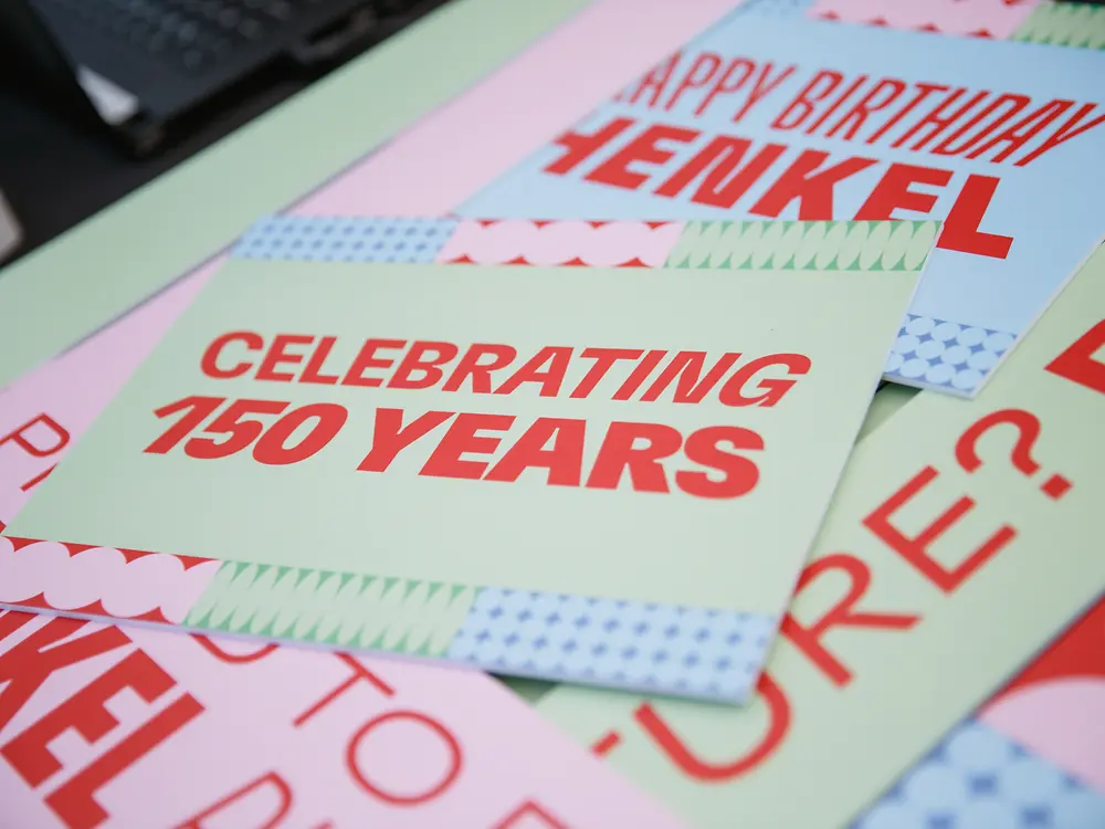 Material gráfico concebido para a celebração do 150.º aniversário da Henkel, com cartões contendo mensagens como “Celebrating 150 Years” e “Happy Birthday Henkel”, sobre uma superfície em tons claros.